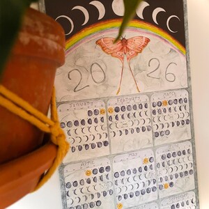 Puede incluir: Calendario de acuarela para 2026 con una ilustraci&oacute;n de polilla, un arco&iacute;ris y fases lunares. El calendario muestra los meses de enero a septiembre, cada uno mostrando las fases de la luna. El calendario est&aacute; dise&ntilde;ado con una parte superior de madera.