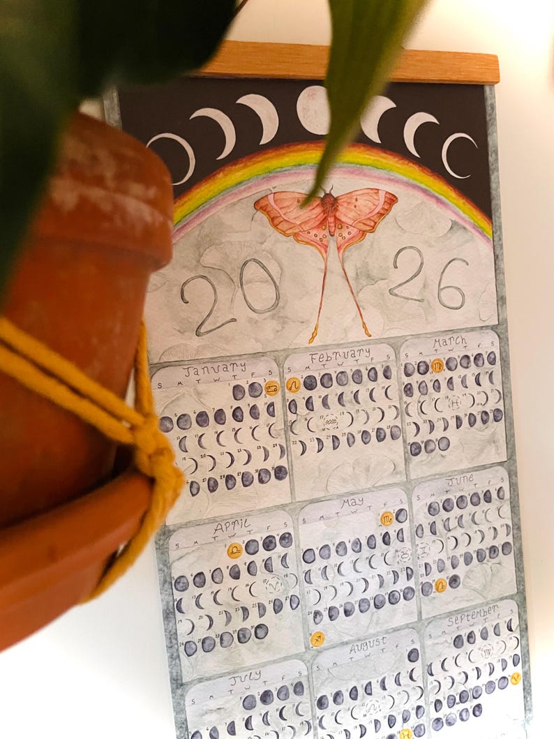 Puede incluir: Calendario de acuarela para 2026 con una ilustraci&oacute;n de polilla, un arco&iacute;ris y fases lunares. El calendario muestra los meses de enero a septiembre, cada uno mostrando las fases de la luna. El calendario est&aacute; dise&ntilde;ado con una parte superior de madera.