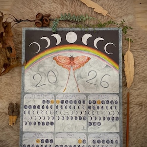 Puede incluir: Calendario de acuarela con una polilla, un arcoíris y fases lunares. El calendario está fechado en 2026 e incluye ilustraciones mensuales de los ciclos lunares. La obra de arte está sobre un fondo oscuro con un borde gris.