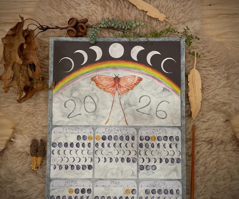 Puede incluir: Calendario de acuarela con una polilla, un arco&iacute;ris y fases lunares. El calendario est&aacute; fechado en 2026 e incluye ilustraciones mensuales de los ciclos lunares. La obra de arte est&aacute; sobre un fondo oscuro con un borde gris.