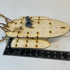 May include: A wooden model spaceship with a decorative design, including a compass rose. The model is approximately 10 inches long. A small figurine is placed on the spaceship. The model is made of light-colored wood.