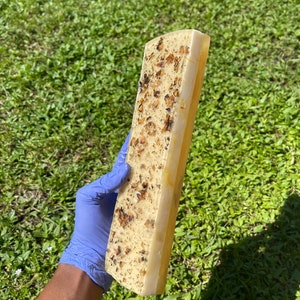 May include: A long, rectangular bar of soap with a pale yellow colour and flecks of brown throughout. The soap is being held in a blue latex glove.