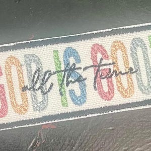 May include: Rectangular patch with the phrase "GOD IS GOOD all the time" in colorful, embroidered letters. The letters are in pastel colors, with a black cursive font for "all the time". The patch has a white background and a black border.