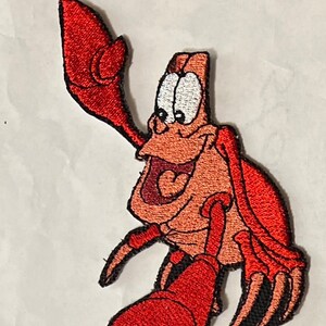 May include: A red and orange embroidered patch of Sebastian the crab from Disney's The Little Mermaid. Sebastian is smiling with his eyes wide open and his claws raised.