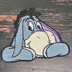 May include: Embroidered patch featuring Eeyore from Winnie the Pooh. The patch has a light blue body, a tan face, and a black mane. The ears are purple, and the eyes are white with black pupils. The patch is designed to be ironed on.