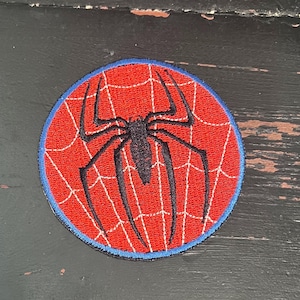 May include: A round, embroidered patch featuring a red background with a white spiderweb design. A black spider is centered on the web. The patch has a blue border. This patch is suitable for clothing or accessories.