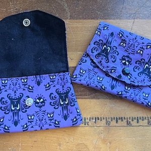 May include: A purple fabric coin purse with a black and white Haunted Mansion pattern. The purse has a flap with a snap closure and a black fabric lining.