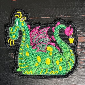 May include: Embroidered patch featuring a whimsical green dragon with yellow spots and accents. The dragon has pink wings and a yellow lantern. The patch is outlined in black.