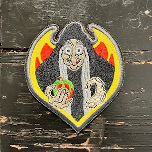 May include: Embroidered patch featuring a cartoon witch holding a green and red apple. The witch has a black hood, gray hair, and a yellow and red background. The patch is outlined in gray and black.