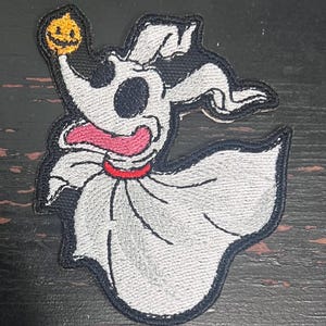 May include: Embroidered patch featuring Zero, the ghost dog from The Nightmare Before Christmas. The patch is white, black, and red, with Zero holding a jack-o'-lantern. The patch has a black border and is likely for clothing or accessories.