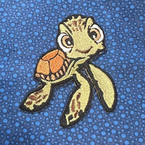 May include: Embroidered patch featuring a cartoon sea turtle with a smiling face, brown shell, and green flippers. The patch has a black border and is set against a blue fabric background with a polka dot pattern.