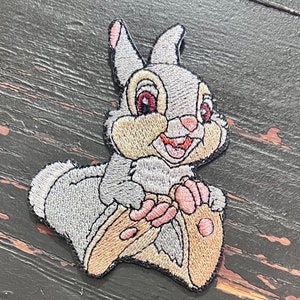 May include: Embroidered iron-on patch featuring a cartoon rabbit with brown fur, pink ears, and red eyes. The rabbit is sitting on a brown log.
