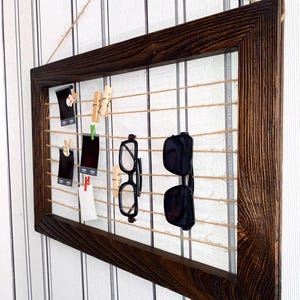 May include: A dark brown wooden frame with twine strung across the inside.  The twine is used to hold photos, postcards, and glasses.  There are several wooden clothespins attached to the twine.