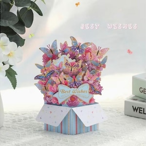 May include: A pop-up card with a butterfly bouquet in pink, blue, and yellow hues. The card is presented in a blue and white striped box, with the words "Best Wishes" on a banner.