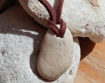 May include: A light brown stone pendant on a brown leather cord. The pendant is resting on a pile of white rocks.