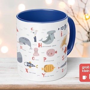 Pode incluir: Caneca de cerâmica branca com interior e alça azuis, com um alfabeto ilustrado de criaturas marinhas. O design inclui um narval, um tubarão-martelo, um polvo e um peixe-cirurgião amarelo. A caneca tem o texto "groß & klein" na lateral.