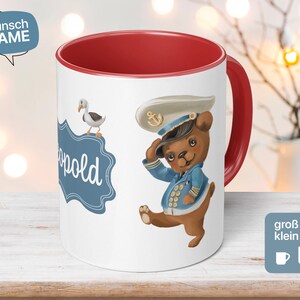 Peut inclure: Une tasse en céramique blanche avec un bord et une poignée rouges. La tasse présente une illustration de dessin animé d'un ours portant un chapeau de marin et tenant un gouvernail de navire. L'ours se tient sur une pancarte bleue et blanche qui dit "Léopold". Une mouette blanche est perchée sur la pancarte. La tasse est sur une surface en bois blanc.
