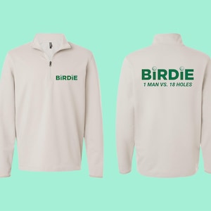 Puede incluir: Un jersey de golf beige claro con cuello con cremallera. La parte delantera muestra la palabra "BIRDIE" en verde, y la parte trasera muestra "BIRDIE" con gráficos de pelotas de golf y el texto "1 MAN VS. 18 HOLES" en verde. Para golfistas.