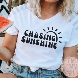 May include: A white t-shirt with black text that reads "Chasing Sunshine" and a stylised sun graphic. The model is wearing the shirt and has a straw hat and sunglasses on.