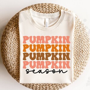 May include: A white t-shirt with the text "Pumpkin season" in a retro font. The text is in shades of orange, brown, and pink.