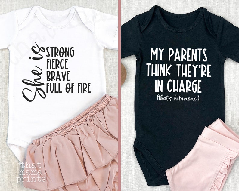 May include: Two baby onesies, one white with black text that reads "She is strong fierce brave full of fire" and one black with white text that reads "My parents think they're in charge (that's hilarious)"