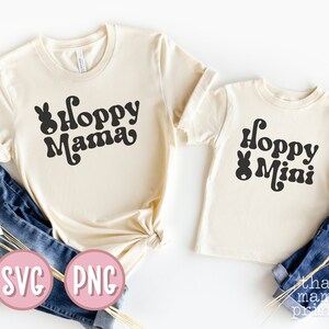 May include: Two cream colored t-shirts with black text and a bunny graphic. The larger shirt says "Hoppy Mama" and the smaller shirt says "Hoppy Mini".