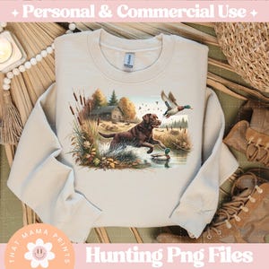 Puede incluir: Una sudadera beige con una escena de caza que presenta un labrador retriever marr&oacute;n y negro, un pato y un fondo de bosque. El texto "That Mama Prints" est&aacute; en la esquina inferior izquierda de la sudadera.