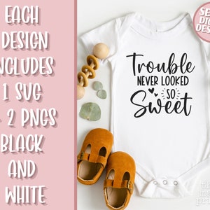 May include: White baby onesie with black text that reads "Trouble never looked so sweet". The onesie is on a white background with brown shoes and green leaves.