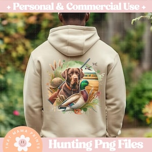 Puede incluir: Una sudadera con capucha beige con un dise&ntilde;o de tem&aacute;tica de caza que presenta un perro marr&oacute;n y negro, un pato verde y una escopeta. El dise&ntilde;o est&aacute; rodeado de vegetaci&oacute;n de estilo acuarela y un fondo marr&oacute;n claro.