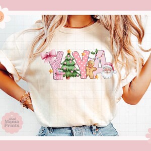 May include: A white t-shirt with a pink and green Christmas-themed design that says "YAYA". The design features a Christmas tree, a gingerbread man, a Santa Claus, and a leopard print pattern.