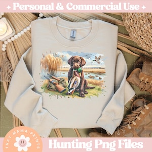 Puede incluir: Una sudadera beige con un estampado gr&aacute;fico de un perro marr&oacute;n y negro con un pato en la boca. El perro est&aacute; de pie en una zona cubierta de hierba con &aacute;rboles y agua en el fondo. El texto "That Mama Prints" est&aacute; en la esquina inferior izquierda de la sudadera.