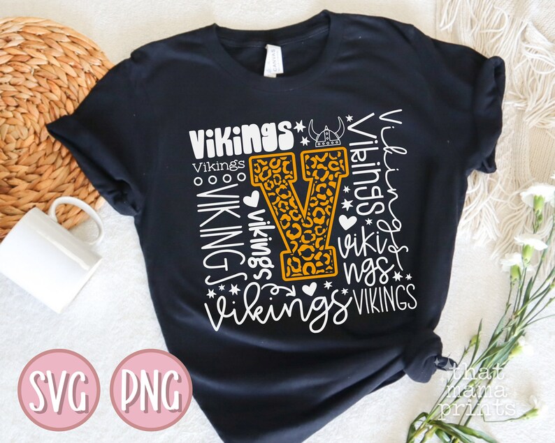 May include: Black t-shirt with a white graphic design of the letter "V" with a leopard print pattern. The graphic is surrounded by the word "Vikings" repeated in different sizes and styles.