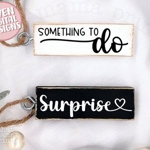 May include: Two wooden signs with twine hangers. The first sign is white with black text that reads "Something to do". The second sign is black with white text that reads "Surprise" with a small heart symbol.