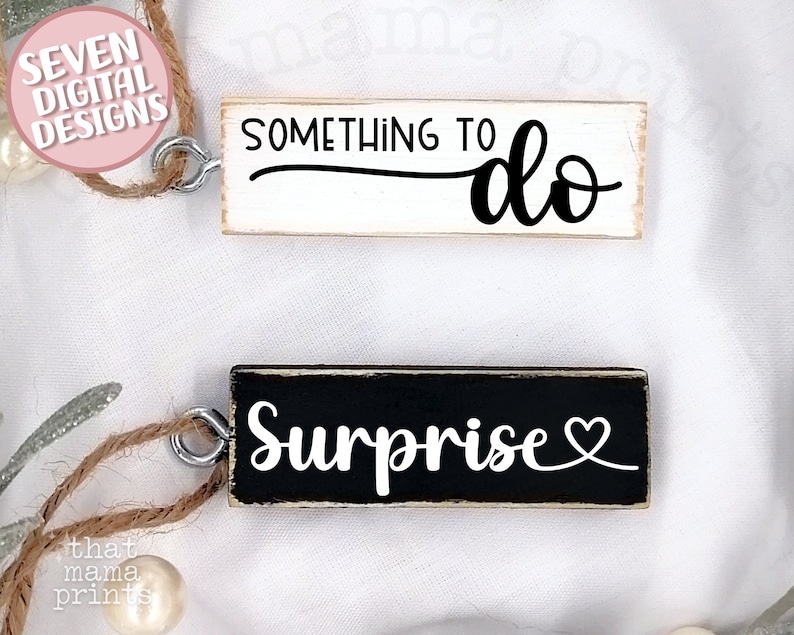 May include: Two wooden signs with twine hangers. The first sign is white with black text that reads "Something to do". The second sign is black with white text that reads "Surprise" with a small heart symbol.