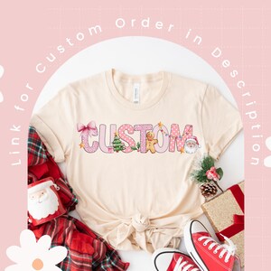May include: A beige t-shirt with a pink and white "CUSTOM" design featuring Christmas-themed elements like a gingerbread man, a Christmas tree, and a Santa Claus. The design is surrounded by pink polka dots and a pink bow.