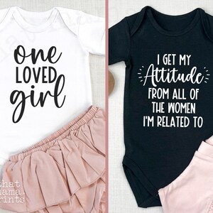 May include: Two baby onesies, one white with black text that reads "one loved girl" and one black with white text that reads "I get my attitude from all of the women I'm related to".