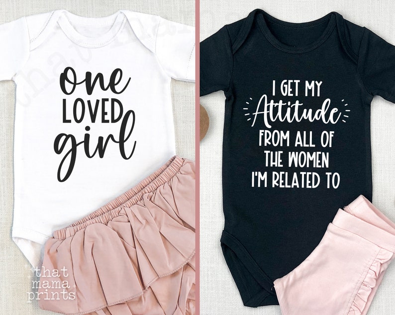 May include: Two baby onesies, one white with black text that reads "one loved girl" and one black with white text that reads "I get my attitude from all of the women I'm related to".