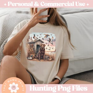 Puede incluir: Una camiseta beige con un estampado gr&aacute;fico de un perro marr&oacute;n y negro de pie en un campo con patos volando por encima. El gr&aacute;fico tambi&eacute;n incluye una peque&ntilde;a caba&ntilde;a de madera y &aacute;rboles. El texto "Hunting Png Files" est&aacute; impreso debajo del gr&aacute;fico.