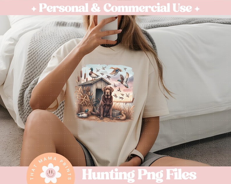 Puede incluir: Una camiseta beige con un estampado gr&aacute;fico de un perro marr&oacute;n y negro de pie en un campo con patos volando por encima. El gr&aacute;fico tambi&eacute;n incluye una peque&ntilde;a caba&ntilde;a de madera y &aacute;rboles. El texto "Hunting Png Files" est&aacute; impreso debajo del gr&aacute;fico.