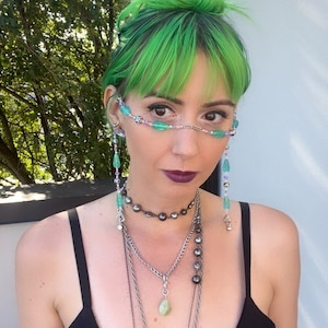 May include: A person with bright green hair wearing a black tank top and multiple necklaces. They are wearing a beaded chain across their face, with green and blue beads.