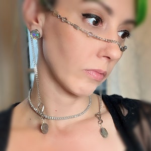 May include: A silver chain nose ring with beads and a silver chain necklace with two silver charms. The necklace is layered over a white choker.