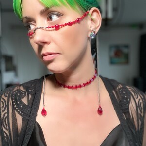 May include: A silver chain with red beads and teardrop-shaped charms hangs from the ears and neck of a person with green hair. The chain is worn as a necklace and a pair of glasses.