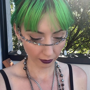 May include: A person with bright green hair wearing a silver chain necklace with a pendant and a silver chain with large black beads. They are also wearing a silver chain with small, iridescent beads across their forehead.