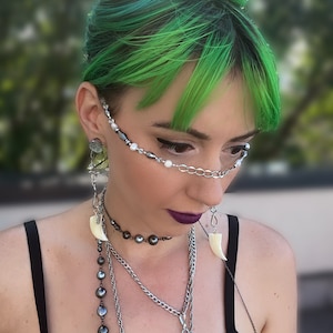 May include: A person with bright green hair is wearing a variety of necklaces, including a choker with dark grey pearls, a silver chain with a green stone pendant, and a long chain with a white bone pendant. The person is also wearing a silver chain with white pearls and dark grey beads across their face.