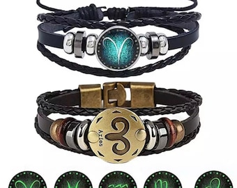 Può includere: Due braccialetti in pelle con chiusure regolabili. Il braccialetto superiore ha un cinturino in pelle nera con un accento di perline argentate e nere e un ciondolo rotondo in vetro con un simbolo zodiacale blu e verde per l'Ariete. Il braccialetto inferiore ha un cinturino in pelle marrone con un accento di perline marroni e argentate e un ciondolo rotondo in metallo con un simbolo zodiacale dorato per l'Ariete. Il testo "Ariete" è scritto sul ciondolo in metallo. Cinque simboli zodiacali aggiuntivi che brillano al buio sono mostrati sotto i braccialetti: Ariete, Pesci, Acquario, Scorpione e Leone.