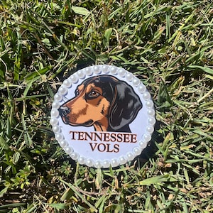 May include: A circular white badge featuring a graphic illustration of a brown and black hound dog head. Below the dog, the text TENNESSEE VOLS is printed in brown capital letters. The edge of the circle is decorated with a border of white pearls.