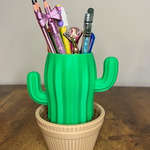 May include: A green cactus-shaped desk organizer filled with colorful pens and pencils. The organizer sits in a beige pot filled with small, brown pebbles. The pencils have pink bows and the pens have diamond-shaped tops.