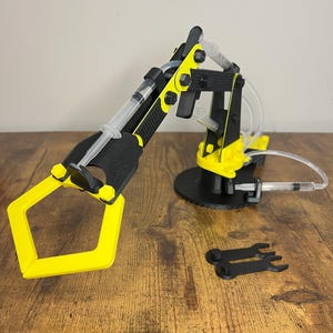 May include: A black and yellow robotic arm with syringes and tubing, resting on a black circular base. Two small black wrenches are also present. The arm's claw is yellow, and the arm is made of plastic.