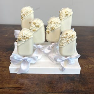 May include: Six white cake pops decorated with gold sprinkles and white frosting on a white stand with white ribbon bows.