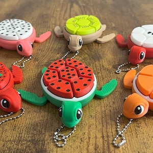 May include: Six colorful turtle keychains with different fruit designs. The turtles are made of soft, squishy material and have a keychain loop attached. The fruit designs include watermelon, strawberry, kiwi, pineapple, and two different types of melon.